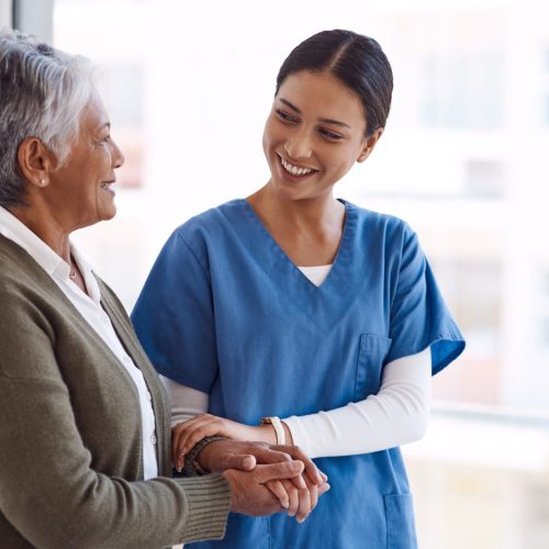 Support, caregiver with senior woman and holding hands for care indoors. Retirement, consulting and professional female nurse with elderly person smiling together for healthcare at nursing home.